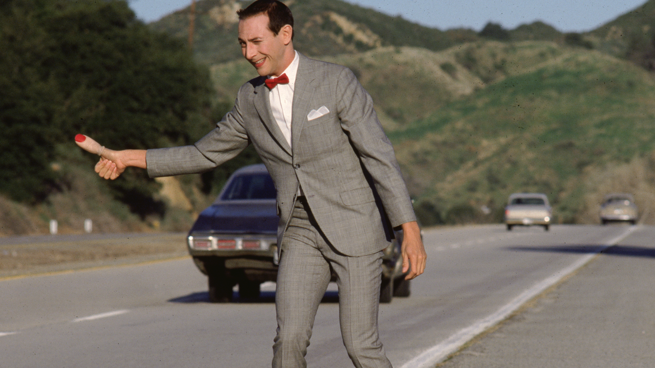Paul Reubens is Pee-wee hitching on the side of the road with a really big thumb in Pee-wee's Big Adventure (1985).